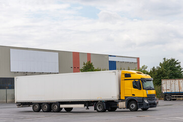 A large semi-trailer with a yellow cab stands near an industrial warehouse.