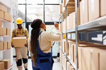 Industrial warehouse employee managing freight inventory, standing near shelf full of cartons. African american woman counting parcels and pointing with pen in shipment storehouse