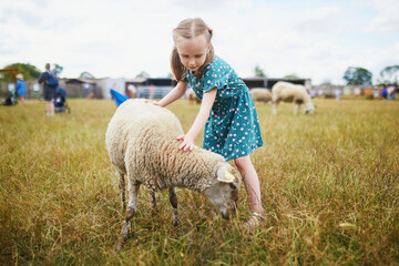 Adorable preschooler girl playing with sheep at farm