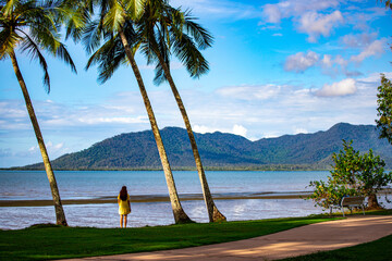 pretty girl in yellow dress admiring  the view of hinchinbrook island from the beach in cardwell, north queensland, australia; tropical beach with palm trees and famous island in the background