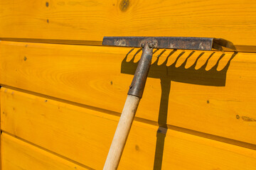 An old rusty rake is leaning against a yellow wooden wall and a contrasting shadow and copy space. ...