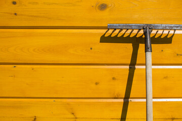 An old rusty rake is leaning against a yellow wooden wall and a contrasting shadow and copy space. ...