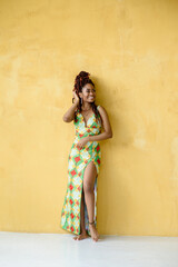 The attractive African woman joyfully smiling while looking away, wearing a long multicolor ornament high slit dress, standing barefoot in front of a yellow wall background with copy space.