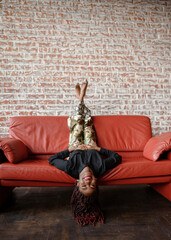 Cheerful young black woman lying upside down on the red leather sofa, looking at the camera and smiling in the room with a brick wall. Concept of joy and happiness. Having fun acting as a child