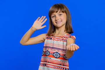 Young cute brunette school girl smiling friendly at camera and waving hands gesturing hello or goodbye, welcoming with hospitable expression. Preteen female child kid isolated on blue background