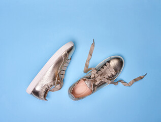 Stylish golden metallic sneakers with wrinkled leather details and white rubber soles on a soft blue background. One shoe has untied shoelaces. The concept of modern stylish footwear