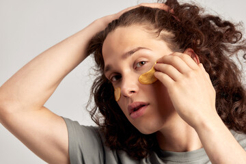 Young handsome long-haired guy applying under eye hydrogel patches on his face. Caucasian brown-haired millennial man practices skin care routine to keep healthy and youthful looking. Studio portrait.