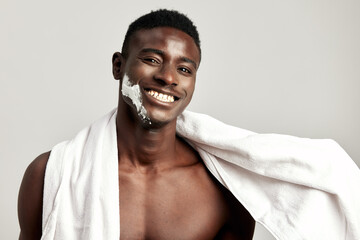 Studio portrait of handsome smiling black guy with naked torso, towel on his shoulder and cream on his face. Afro American man uses beauty products in his skincare routine to maintain healthy look.