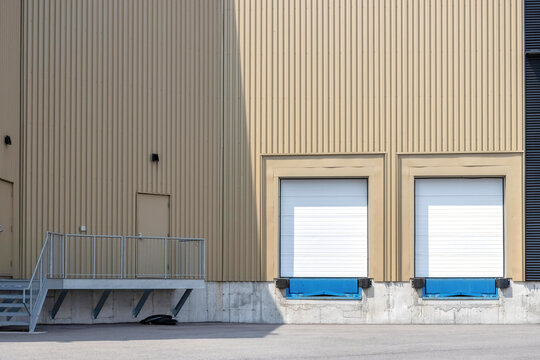 Facade of a high bay commercial building showing empty loading docks with closed metal doors, vertical corrugated metal siding, steel stair to entry doorway, daytime, sunny, nobody