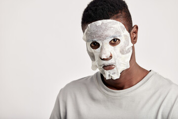 Close-up studio portrait of young black man with moisturizing paper mask on his face. Happy African American millennial man uses beauty products in his skincare routine to maintain healthy look.