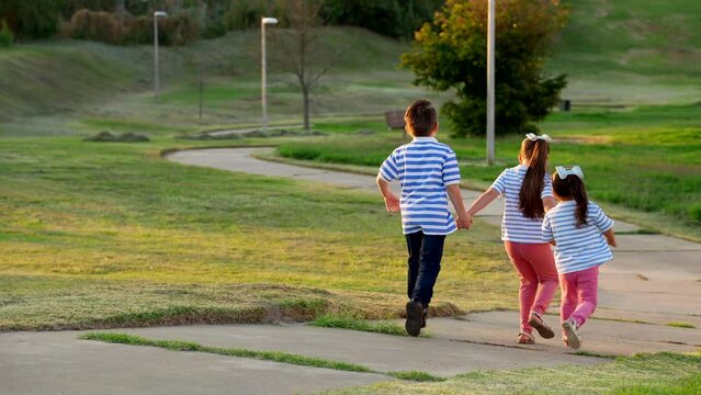 Tres ni&ntilde;os hermanos amigos latinos mexicanos corriendo bajando por el sendero en un parque al exterior al aire libre jugando y disfrutando sonrientes en un hermoso atardecer juntos tomados de la mano 