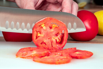 CLOSE UP OF CHEF KNIFE CUTTING TOMATOES ON THE CHOPPING BOARD COOKING A RECIPE IN THE KITCHEN.