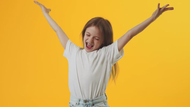 Funny Caucasian girl kid wearing white t-shirt hold fan of cash money in dollar banknotes and throws money in all directions on plain yellow background in studio