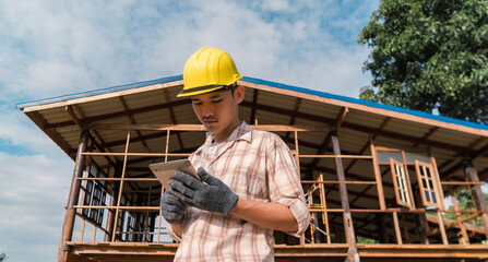 Naklejka na ściany i meble Asian carpenter holding tablet working at construction site using technology Wear a helmet and goggles to prevent dust.
