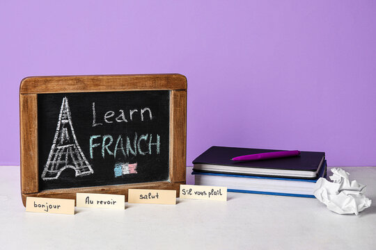 Chalkboard with text LEARN FRENCH, words and books on table near lilac wall