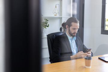 Young caucasian handsome businessman with beard using smartphone sitting office table. Personal mobile communication. Man send message or search browsing information. Ordering work via online system.