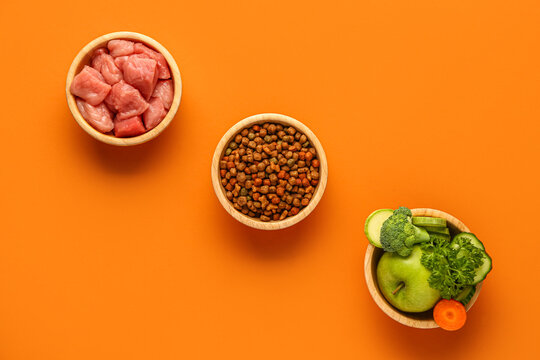 Bowls with dry pet food, raw meat and natural products on orange background