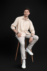 Handsome happy young man with wristwatch sitting on chair against black background