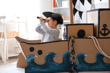 Cute little pirate playing with spyglass in cardboard ship at home