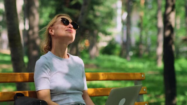 Relaxed blonde woman sitting on the bench with a laptop, enjoying the beautiful weather and fresh air. Female freelancer working remotely in the summer city park.
