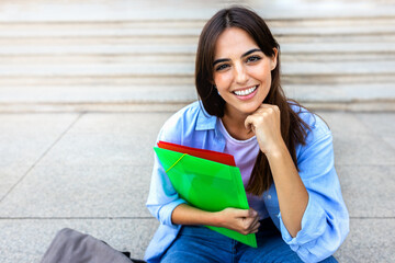 Young beautiful female college student sitting on stairs outside university building looking at camera. Copy space.