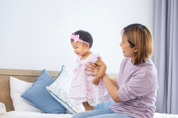 Asian mother is holding her pretty smiling baby daughter while spending quality time in the bed for family happiness and parenting