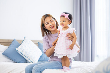 Obraz na płótnie Canvas Asian mother is holding her pretty smiling baby daughter while spending quality time in the bed for family happiness and parenting