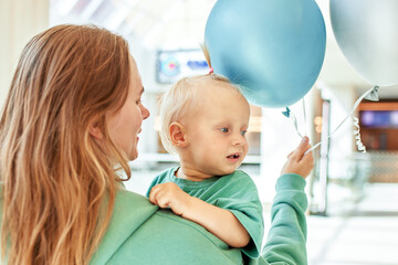 Close-up portrait of mother with a baby in her arms and colored balloons. Young pretty mom with a little daughter in the same green clothes are walking around the mall and having fun. Happy family.