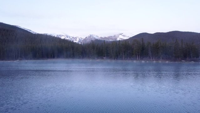 Steam rising off of Colorado mountain lake with snowcapped Rocky Mountains in the background, pan left aerial