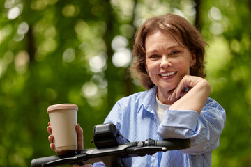 Adorable mature woman with electric scooter and paper coffee cup in the park. Joyful smiling...