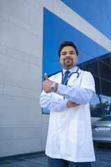 Young indian doctor standing out of hospital and showing thumps up