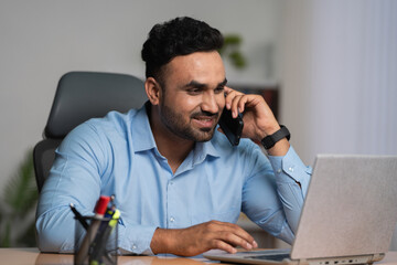 Indian businessman talking on mobile phone while working on laptop at office - concept of financial advisor, multi tasking and Corporate Lifestyle