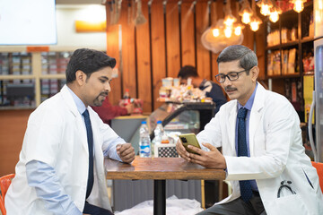 Two indian doctors sitting hospital canteen looking in smartphone and discussing.