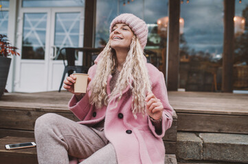 fashionable mature woman in pink knitted beanie hat and stylish coat sits outside in cafe and drinks coffee from disposable paper cup. Middle aged woman positive and smiling with pigtails in the city 