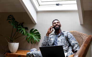 African american male soldier wearing military uniform with laptop and talking on smartphone at home