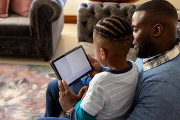 Happy african american father and son sitting in living room at home using tablet with copy space