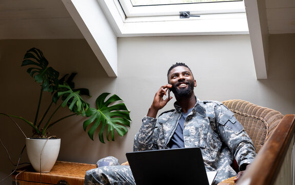 African american male soldier wearing military uniform with laptop and talking on smartphone at home