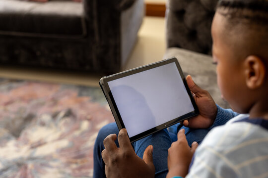 African american son sitting in living room at home with father using tablet with copy space