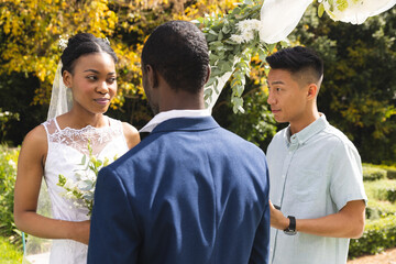 Happy diverse male officiant with bride and groom at outdoor wedding ceremony in sunny garden - Powered by Adobe