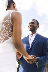Happy african american bride and groom holding hands at wedding ceremony in sunny garden