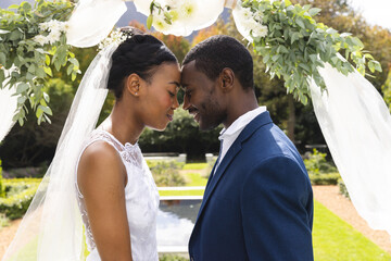 Happy african american bride and groom touching heads under wedding arch at ceremony in sunny garden