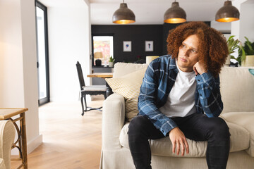 Thoughtful, sad biracial man sitting on couch in living room, looking away, copy space - Powered by Adobe