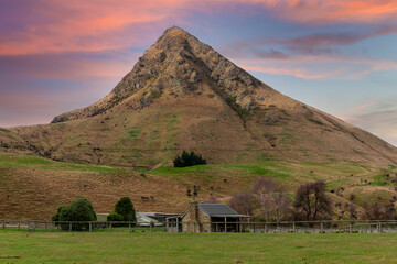 Photograph of an old small stone farm cottage in a lush green agricultural field in front of a large hill on the South Island of New Zealand - Powered by Adobe