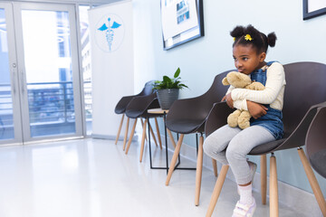 African american girl patient sitting in waiting room, embracing teddy bear at hospital, copy space