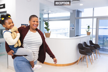 Happy african american mother and daughter walking in waiting room at hospital, copy space