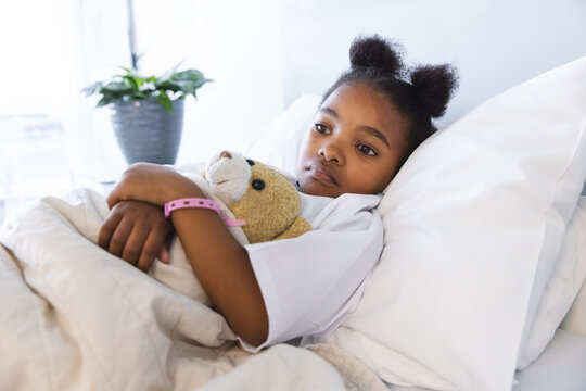 Sad african american girl patient lying on bed with teddy bear in patient room at hospital