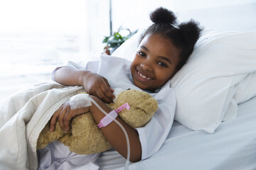 Portrait of happy african american girl patient lying on bed in patient room at hospital - Powered by Adobe