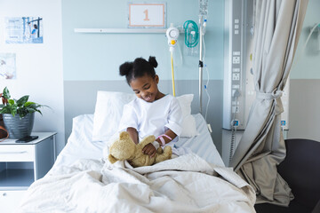 Happy african american girl patient lying on bed playing with teddy bear in patient room at hospital