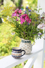 A beautiful ceramic coffee cup and jug with wildflowers on the railing of the summer veranda. Selective focus. 