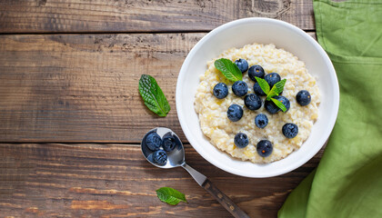 Creamy millet breakfast porridge topped with blueberries and mint in a white bowl on a rustic wooden table, horizontal view from above, flat lay, free space - obrazy, fototapety, plakaty